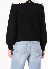 Pull en maille noire avec un col en dentelle blanche ; présente des manches bouffantes, un ourlet côtelé et une fermeture à boutons dans le dos ; associé à un jean bleu clair.