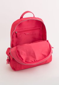Rucsac roșu din material textil, cu partea superioară rotunjită, compartimente cu fermoar dublu și un capac frontal cu închidere magnetică. Textură netedă, design practic.