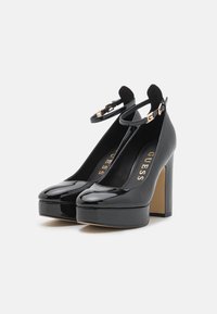 Scarpe con tacco alto in pelle verniciata nera, con piattaforma, punta arrotondata, cinturino alla caviglia e dettaglio della fibbia in tono oro.