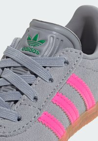 Pilki zomšiniai sportiniai batai su rožinėmis juostelėmis, pilkomis raištelėmis ir žaliu Adidas logotipu ant liežuvėlio. Tekstūruoti, su guminiais padais.