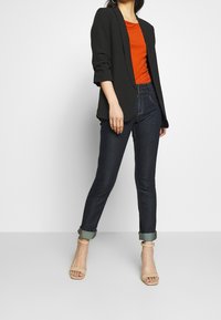 Czarna marynarka na pomarańczowej bluzce, do tego ciemne dżinsy i beżowe sandały na obcasie z paskami. Podwinięte mankiety w dżinsach i dopasowany fason marynarki.