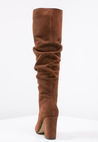 Bottes en daim marron hauteur genou avec un design froncé, un talon épais et une texture lisse. Dotées d'un bout arrondi et de détails de couture minimaux.