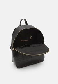 Musta reppu, jossa on kiiltävä krokotiilinäköinen pinta, kultainen vetoketju ja tilava sisustus, jossa on yksi vetoketjullinen tasku ja tasainen lokero.