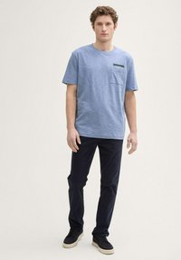 Camiseta a rayas azul con mangas cortas y un bolsillo en el pecho, combinada con pantalones oscuros y zapatillas negras sobre un fondo neutro.