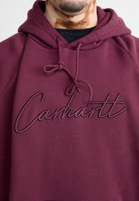 Felpa con cappuccio color mogano caratterizzata da un ricamo testurizzato della parola "Carhartt" nel colore abbinato, con cordini regolabili e una vestibilità comoda.