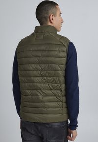 Zeleni puffer brezrokavnik z visokim ovratnikom, prešito obliko in gladko, sijočo teksturo, nosi preko temnomodre majice s dolgimi rokavi.