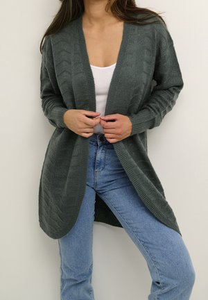 Femme portant un cardigan en maille torsadée gris foncé sur un débardeur blanc et un jean bleu, tenant les bords avant du cardigan.