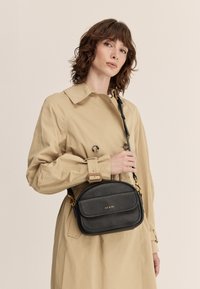 Čierna kožená crossbody taška so zlatými detailmi, zakrivený tvar a dve predné vrecká. Nosená cez béžový trenčkot s gombíkovými detailmi.