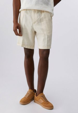 Uomo che indossa pantaloncini di lino beige e sneakers in camoscio marrone chiaro, in piedi davanti a uno sfondo chiaro e semplice.