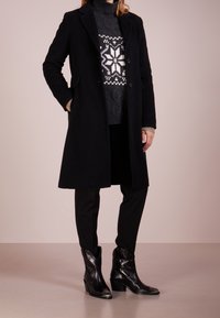 Zwarte wollen jas met twee knopen, met een getailleerd ontwerp. Draag het over een grijze gebreide trui met een sneeuwvlokpatroon en zwarte broek. Zwarte laarzen.