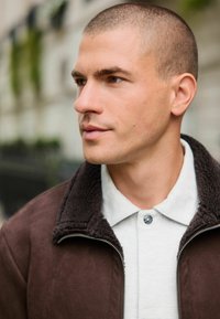 Mann mit kurzem Buzzcut, der eine dunkelbraune Jacke über einem weißen Hemd mit Kragen trägt und nach links schaut, vor einem verschwommenen Hintergrund im Freien.