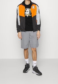 Man i orange och svart sportjacka, svart grafisk T-shirt, grå shorts, vita strumpor och svarta träningsskor som står mot en enfärgad vägg.