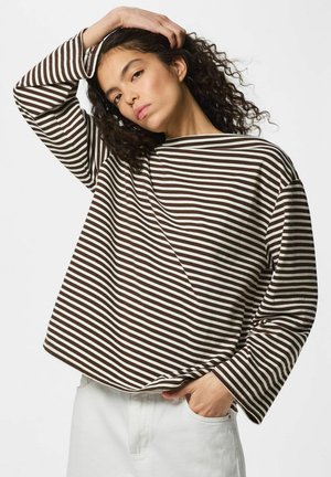 Frau mit lockigem Haar trägt ein braun-weiß gestreiftes Langarmshirt und weiße Hose, eine Hand in der Tasche, die andere berührt ihren Kopf.