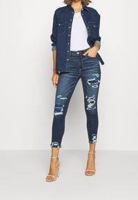 Smala jeans i mörkblå denim med slitdetaljer, fransade fållar och hög midja. Kombineras med en denimskjorta och beige klackar.