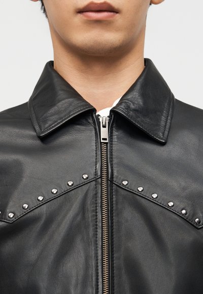 Κοντινό πλάνο ενός ατόμου που φοράει μαύρο δερμάτινο μπουφάν με ασημένιες τρουκς κατά μήκος μιας γραμμής σχήματος βέλους και κεντρικό μεταλλικό φερμουάρ.