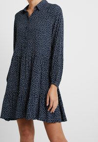 Abito blu navy con colletto, maniche lunghe e design a strati. Presenta un piccolo motivo geometrico bianco su tutta la superficie. Tessuto dalla consistenza morbida.