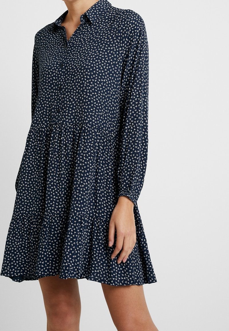 Abito blu navy con colletto, maniche lunghe e design a strati. Presenta un piccolo motivo geometrico bianco su tutta la superficie. Tessuto dalla consistenza morbida.