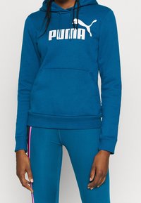 Modrá mikina s kapucí vyrobená z měkké látky, která má klokanní kapsu a bílý logo "PUMA". Kombinováno s odpovídajícími modrými legíny.