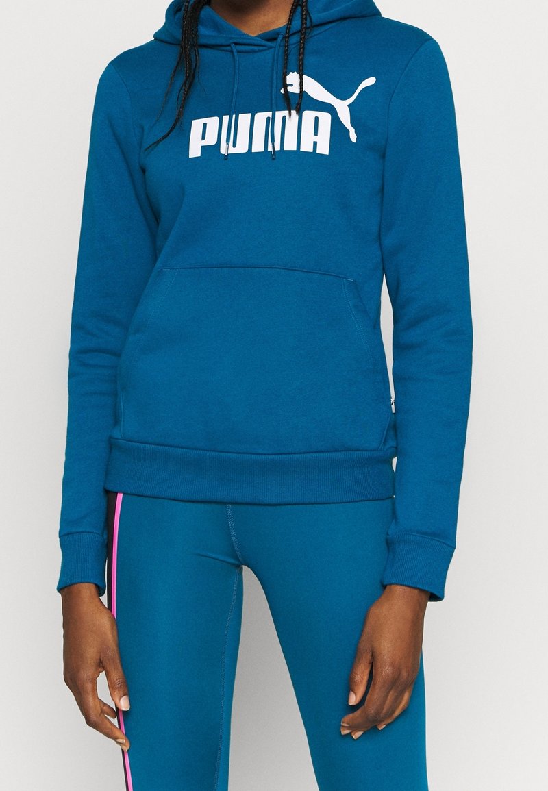 Modrá mikina s kapucí vyrobená z měkké látky, která má klokanní kapsu a bílý logo "PUMA". Kombinováno s odpovídajícími modrými legíny.
