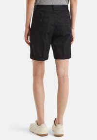 Černé bavlněné šortky s ohrnutými lemy, s dvěma zadními kapsami na knoflíky, zobrazené z pohledu zezadu. Obuv tvoří světlá teniska.