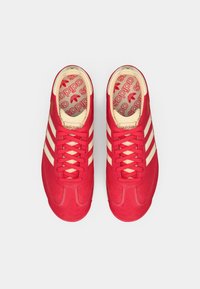 Červené semišové tenisky s krémovými pruhmi a šnúrkami, so štruktúrovanou podrážkou a vnútornou vložkou s opakujúcim sa vzorom loga.