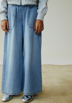 Vaqueros de mezclilla azul claro de pierna ancha con dos bolsillos delanteros, cintura alta y corte recto. Combinados con zapatos metálicos plateados.