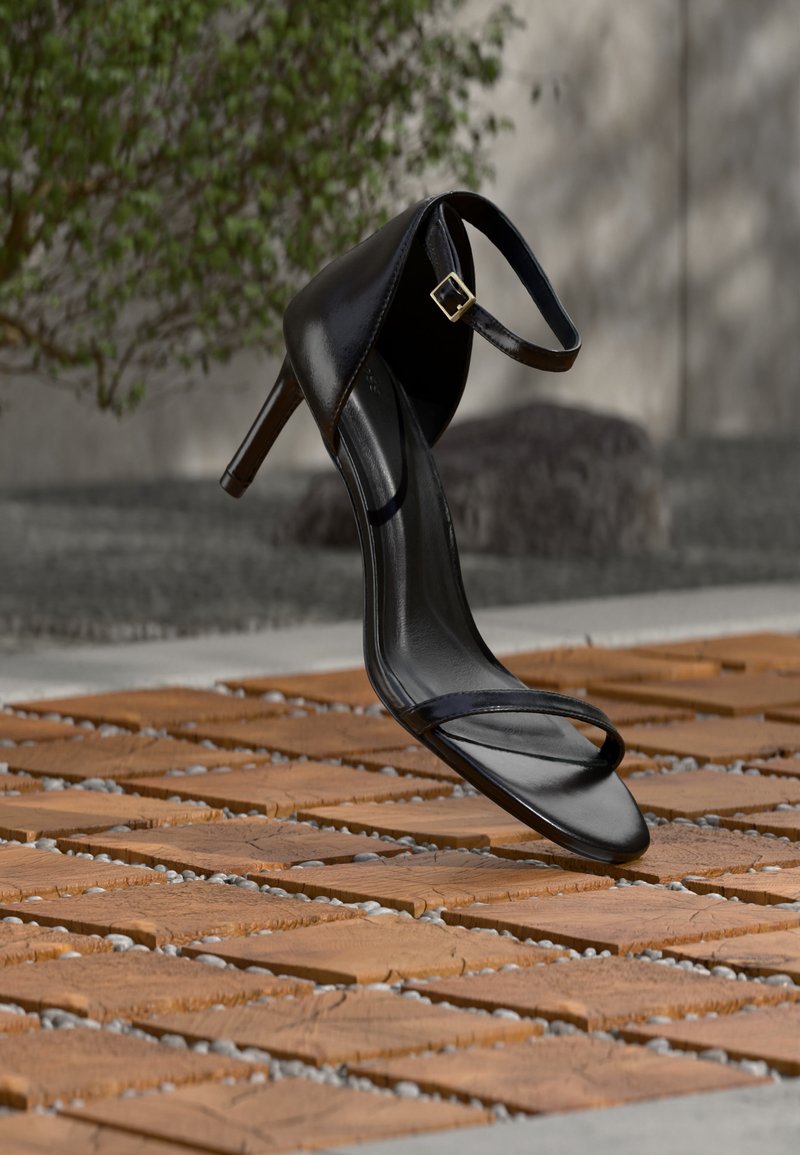 Μαύρο σανδάλι με ψηλό τακούνι και λουράκι στον αστράγαλο, κρεμασμένο πάνω από ξύλινο πλακίδιο δαπέδου σε εξωτερικό χώρο, με θολό φόντο πράσινου και πέτρας.