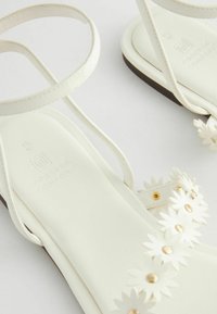 Weiße Sandalen haben ein flaches Design mit dünnen Riemen und floralen Verzierungen. Die Innensohle ist strukturiert und trägt die Aufschrift "Forever Comfort".