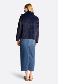 Jachetă din blană sintetică bleumarin cu guler înalt, mâneci lungi și un design elegant, asociată cu o fustă din denim de lungime medie și sandale cu toc din lemn.