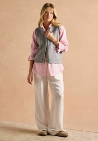 Grauer Wollwesten über einem rosa-weiß gestreiften Hemd, kombiniert mit weißen weiten Hosen und beigen Slippern. Lässiges Ensemble.
