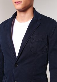 Blazer in cotone blu navy con risvolto classico, chiusura con un unico bottone e taschino sul petto con dettagli di cucitura visibili.