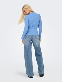 Blondīne sieviete, kas vēršas prom un valkā pieguļošu garu zilu topu un vaļīgas gaiši zilas džinsas ar melniem apaviem uz vienkārša fona.