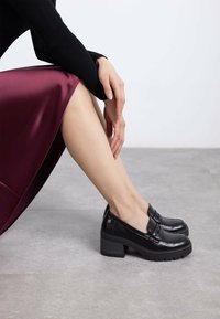 Mocasines de cuero negro con tacón, punta redondeada, plataforma elevada y detalles cosidos. Combinados con una falda de satén burdeos. Textura suave.