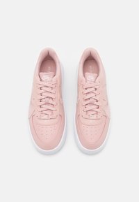 Růžové kožené tenisky s hladkým povrchem, bílé gumové podrážky, perforovaná špička a šněrování. Obsahují logo Nike na jazyku a boku.