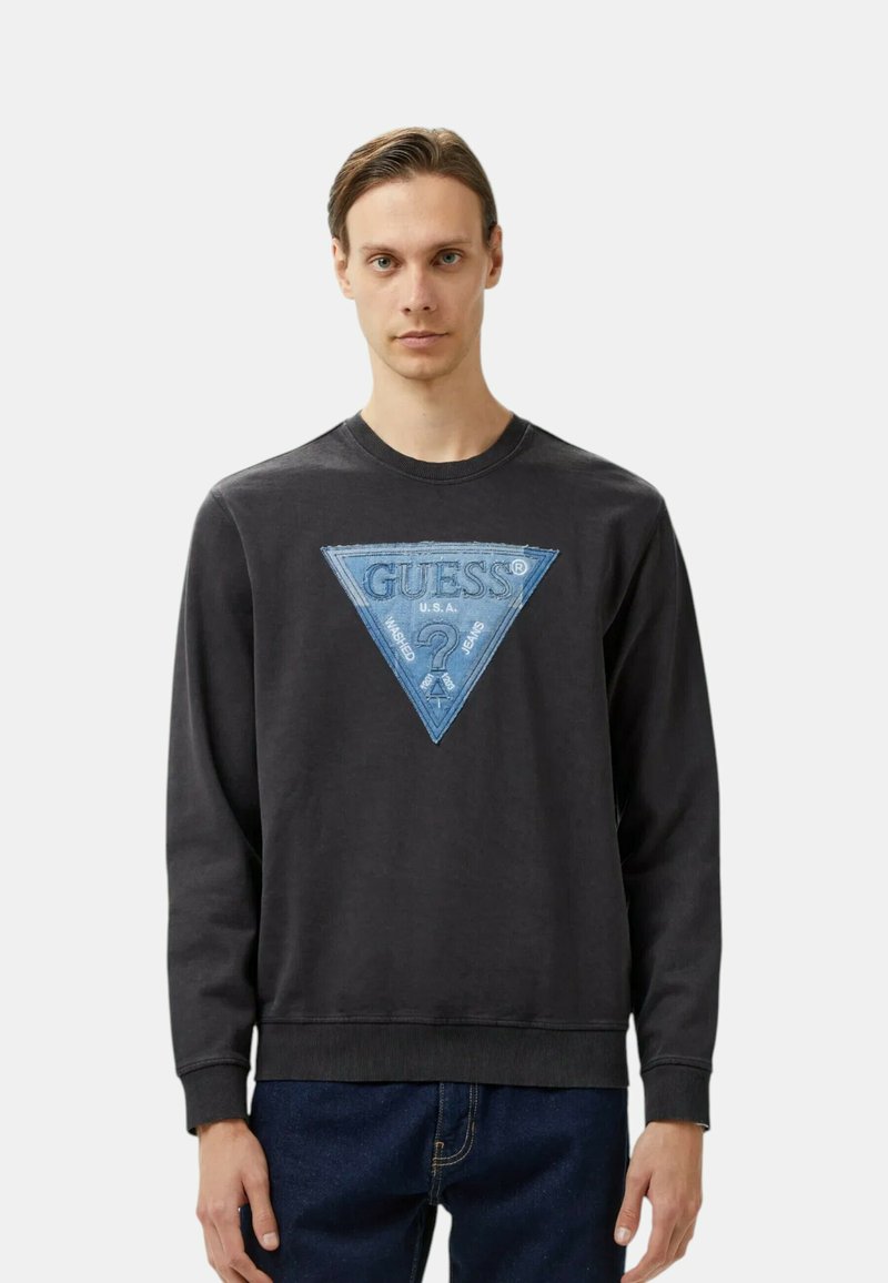 Felpa nera con un logo triangolare in denim blu che presenta il design "GUESS" e un punto interrogativo. Collo semplice, maniche lunghe e polsini a costine.