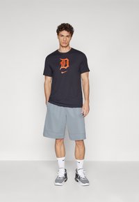 Tamsiai pilkas marškinėliai su oranžiniu logotipu, derinami su šviesiai pilkomis kelnių šortais iki kelių. Modelis vilki baltas kojines ir pilkas sportines batus.