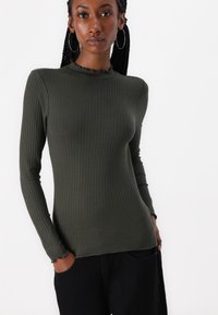 Olive green ribbed long-sleeve top with a ruffled neckline and fitted silhouette, paired with black pants and a relaxed pose.