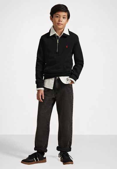 Pullover nero con zip e logo rosso, indossato sopra una camicia bianca, abbinato a jeans larghi scuri e scarpe nere con dettagli bianchi.