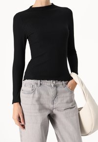 Svart ribbad långärmad topp med hög krage och vågig nederkant, parat med ljusgrå denimjeans och en krämfärgad axelväska.