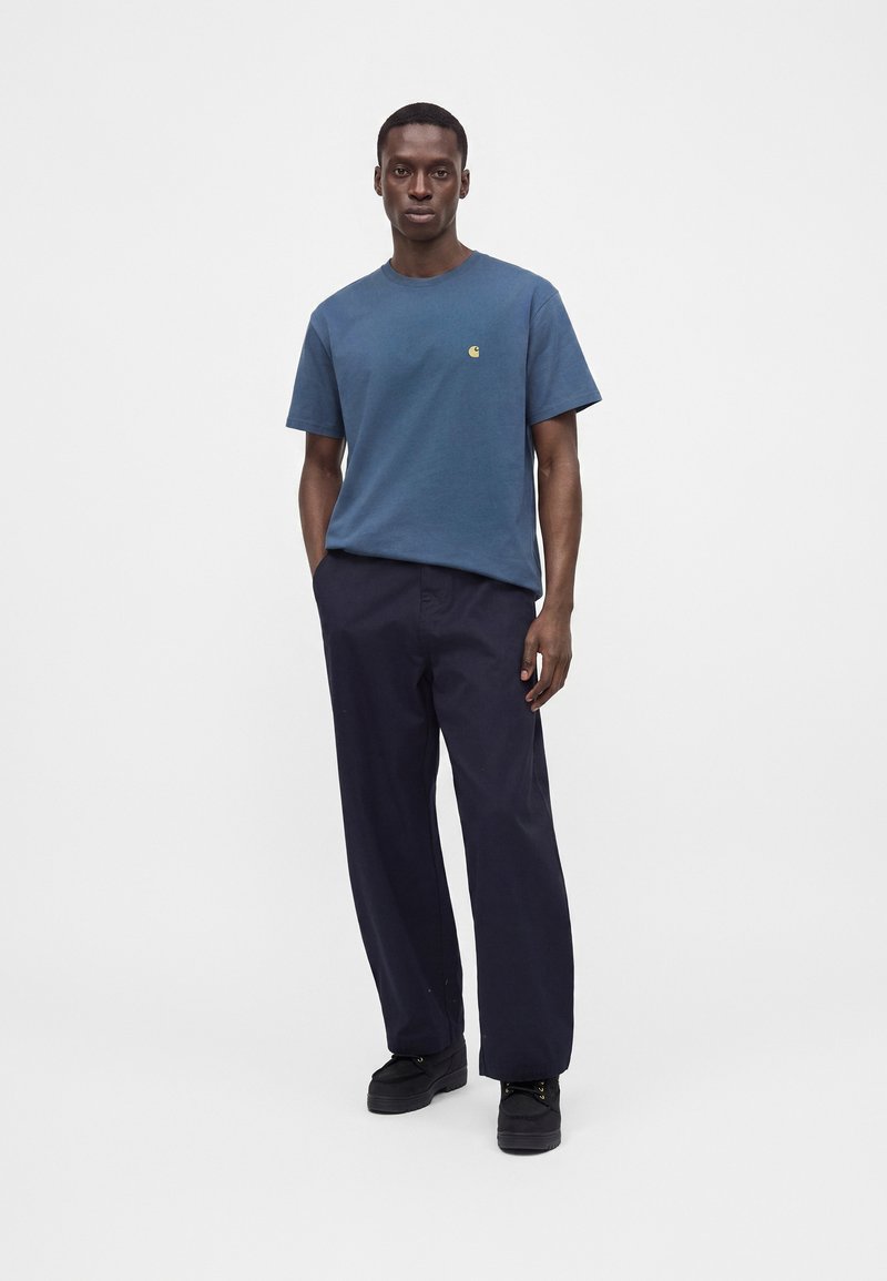Mann steht und trägt ein blaues Kurzarmhemd mit kleinem Logo, dunkle lockere Hose und schwarze Schuhe vor einem schlichten weißen Hintergrund.