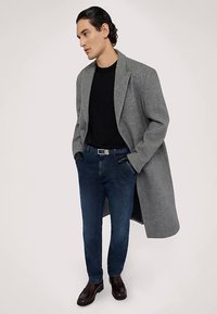 Grå oversized kappa, svart skjorta, mörkblå jeans och mörkbruna skor. Kappan har breda slag och en avslappnad passform.