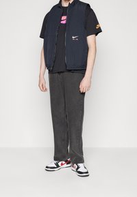 Marinblå ärmlös puffväst, svart T-shirt med rosa logotyp, mörkgrå jeans och svarta sneakers med röda detaljer. Slät textur, avslappnad passform.