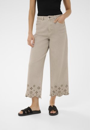 Nő bézs színű, széles szárú nadrágban, virágmintás kivágással a szegélyén, fekete szandálban, kezét a zsebében tartva, egyszerű fehér háttér előtt.