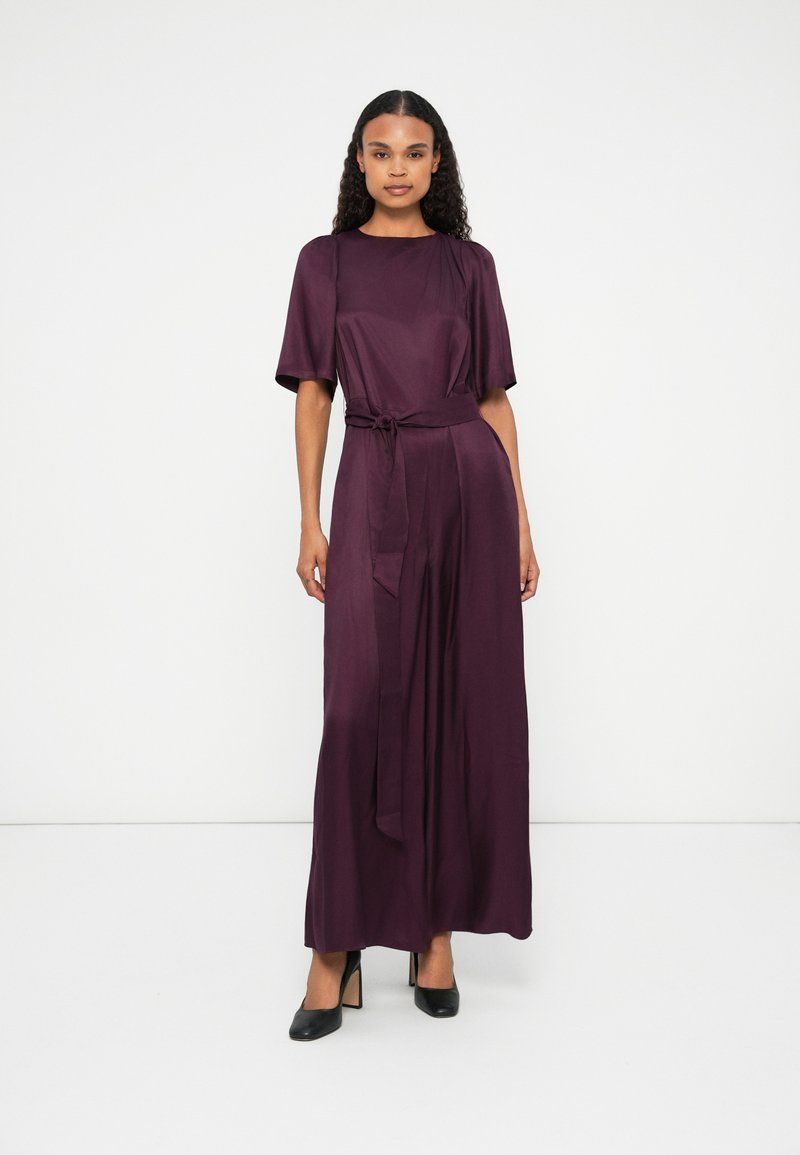 Vestido longo e roxo escuro com mangas curtas, detalhe de cintura amarrada e uma textura suave e fluida. Usado com sapatos de salto alto pretos.