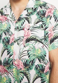 Camicia a maniche corte con sfondo bianco e motivo di fenicotteri rosa vivaci e foglie verdi; include una tasca sul petto e chiusura con bottoni.