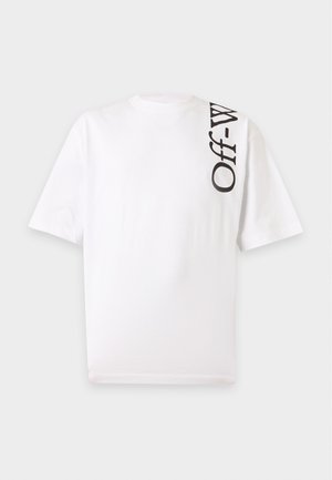 Un homme se tient debout, les mains dans les poches, portant un T-shirt blanc avec un texte vertical "Off-W" et un pantalon noir avec les textes "White™" et "Off", chaussures noires et blanches.