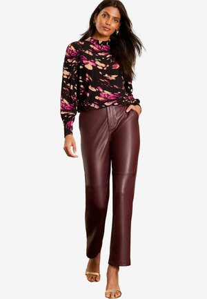 Schwarze Bluse mit lila und rosa abstraktem Muster, kombiniert mit figurschmeichelnden bordeauxfarbenen Kunstlederhosen. Das Model steht vor einem weißen Hintergrund.