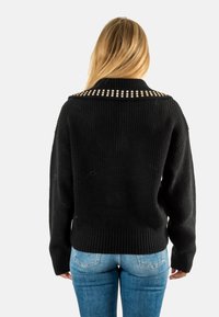 Maglione nero lavorato a maglia con una texture a coste, caratterizzato da un ampio colletto adornato con borchie metalliche. Abbinato a jeans in denim blu.