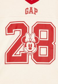 Kremskrāsas T-kreklis ar sarkano V veida kaklu, kam augšpusē ir uzraksts "GAP", skaitlis "28", un centrā sarkana Minnie Mouse grafika.