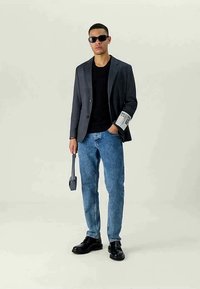 Man wearing dark sunglasses, black shirt, gray blazer with white tag, blue jeans, black shoes, holding a small gray bag.